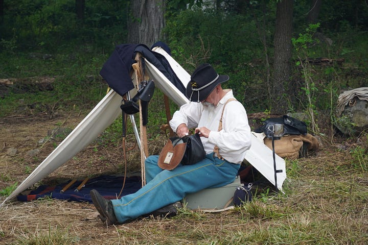 A man in Civil War attire sits in front of a white tent and a man in a gray vest of period attire stands behind a table of medical equipment.