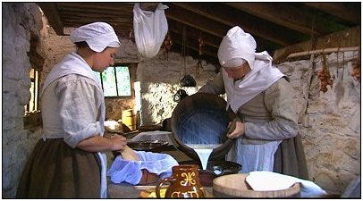 In the first image, two women in early 17th-century clothing work in a historic kitchen. One is pouring milk from a bucket into a bowl. The other is putting her hand into a bowl lined with a cloth. In the second photo, a bearded man in 17th-century clothing leans over two heritage breed pigs in a sunny grove.