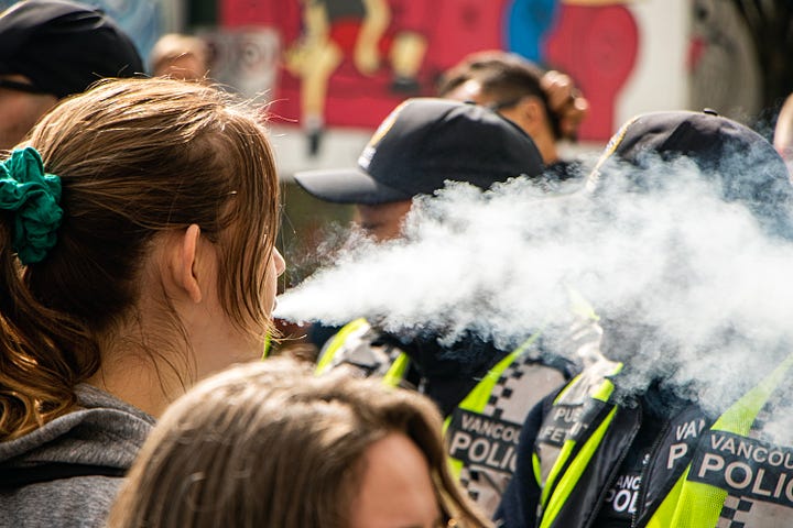 First image: A woman blows a cloud of vapour in the faces of police. A friend who was down there tells me her group was trolling the police the whole time, including taking drags from their vape and blowing it at the police. Second image: A person with a white shirt and a white wrap around their head and face and a black cap holds up a sign that reads: "Cops are better at committing crimes than solving them." Third image: An Indigenous person, back facing the line of police, is holding a bowl of smoking sage. After holding a cap overtop of the sage smoke, they turned the cap over, and smoke can be seen trickling upwards, out of the hat. Fourth image: A couple of police officers are seen at the front line. One is wearing a mask and looking dead ahead. The other is looking at something to the left of the camera's perspective.