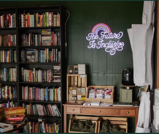 a neon, pastel sign with a rainbow and cursive writing, hung above a table with merchandise, says "The Future is Inclusive." A yellow chair and couch encircle a coffee table with a plant on top, creating an inviting gathering space..