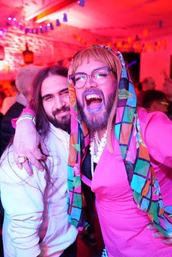 A long-haired scruffy guy and a colorful non-binary person in a bright pink dress and blue lipstick embrace at Feliza Gay and Lesbian Club. A drag queen dressed in fire negine red with a long trailing blonde braid poses at Work Bar.