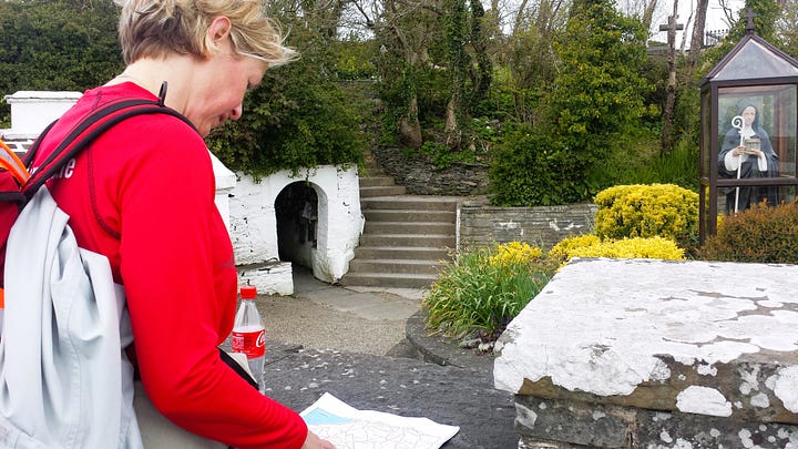 1. Holy well with stone wall and central entrance to access water, tall hedge behind the well. 2. Stone wall surrounding well with steps down to access water, twisted hawthorn tree beside it to left. 3. Paved path leading to well entrance to water underground. 4. Whitewashed entrance to well with steps to left, woman in red top sitting on wall to right looking at a map.