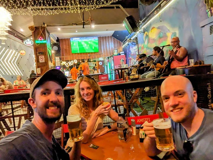 A church in São Paulo, one traveler smiling next to a man's portrait, two travelers waiting for the subway, and all three travelers smiling in a bar and toasting beers