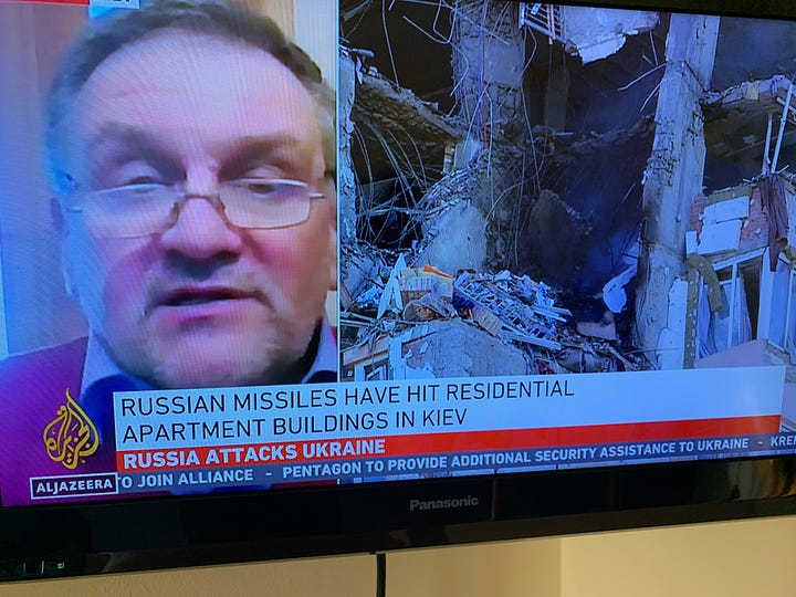 tv news screen grab showing building with bomb damage, refugee help point at train staition. Sign reading "Help for Ukraine here" and a chart of phrases in different languages.