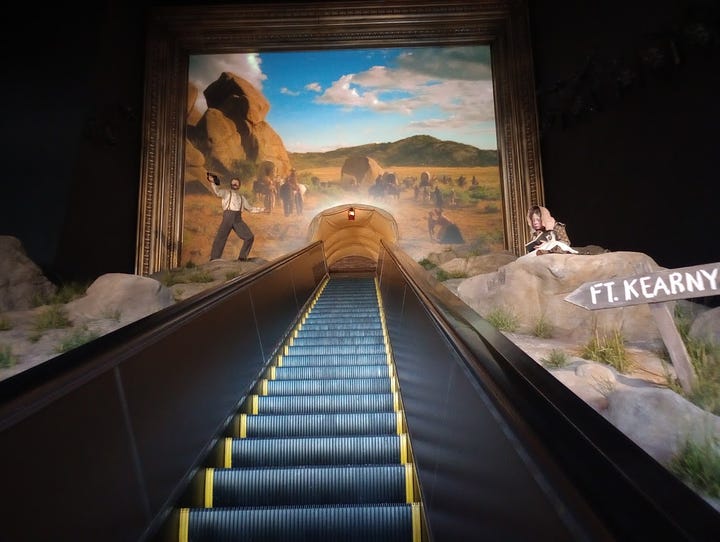 Long elevator with tableau of dry sand and rocks on either side, and a desert view of wagons at top. Indian woman in traditional dress with non-Native man in beard, buckskin, and carrying a rifle, man in wide-brimmed hat clambering up rocky hillside, followed by woman in dress and bonnet. Close view from top of escalator reveals that the passage at top imitates walking through a covered wagonn