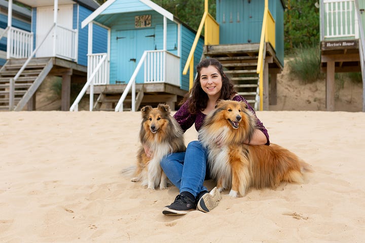 Camilla Fellas Arnold holds a copy of her book Unravelling Inwards. Camilla sits with her two Shetland sheepdogs in front of some colourful beach huts.