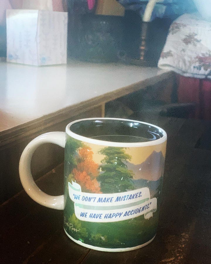 A mug, on one side a photo of Bob Ross with a paintbrush in hand, on the other, trees and and a landscape with the saying, "We don't make mistakes. We have happy accidents."