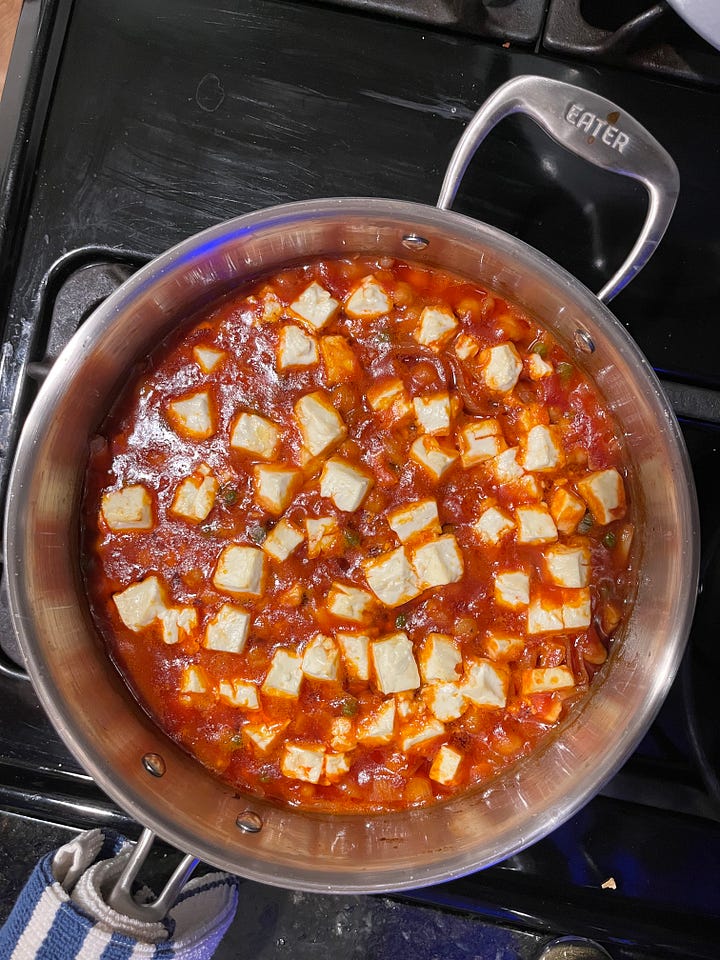 A photo of cookbooks on a shelf with Post-It tabs sticking up from the pages; a view of an onigiri mold with rice and salmon inside; a table with a pot of kabsa, a bowl of greens, and a small bowl of yogurt; a view of a pan with cooked harissa chickpeas and pieces of feta.