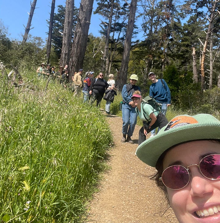 The left image shows QBBA co-leads Paige Pritchard and Alyssa Winn wearing purple and blue hats, cheekily embroidered with the phrase "Check it out, I'm gay." On the right, a selfie taken by Paige shows a group of seven birders on a hard-pack gravel trail through what appears to be a cedar or pine-filled wooded area.