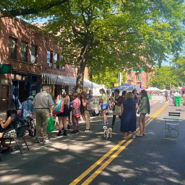 On the left, a group of people crowds around a vendor on the open street. On the right, people walk by tables covered in white tents.