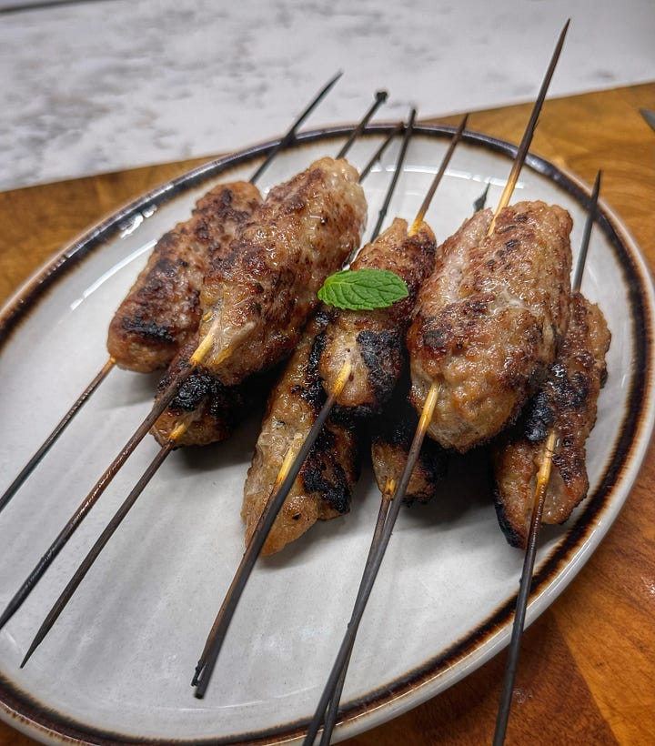 One photo of the skewered sausages cooking on a metal flat-top griddle over a charcoal grill. And a photo of the finished, browned skewered sausages piled on a plate, topped with a single, very small mint leaf. 