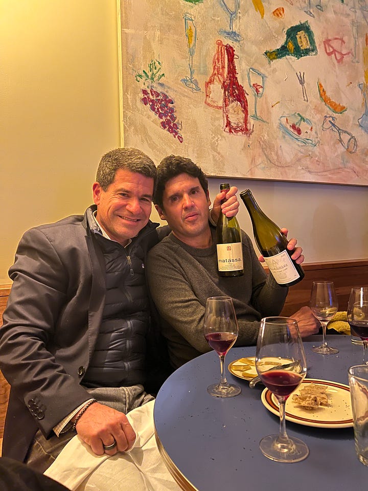 Glass of red wine on a blue table with a couple bottles of wine in the background. second picture: Two men holding sitting at a table, each holding a bottle of wine with wine glasses and food on the table in front of them.  