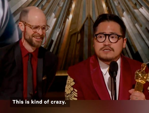 Daniel Kwan at the microphone holding an Oscar with Daniel Scheinert at his side watching
