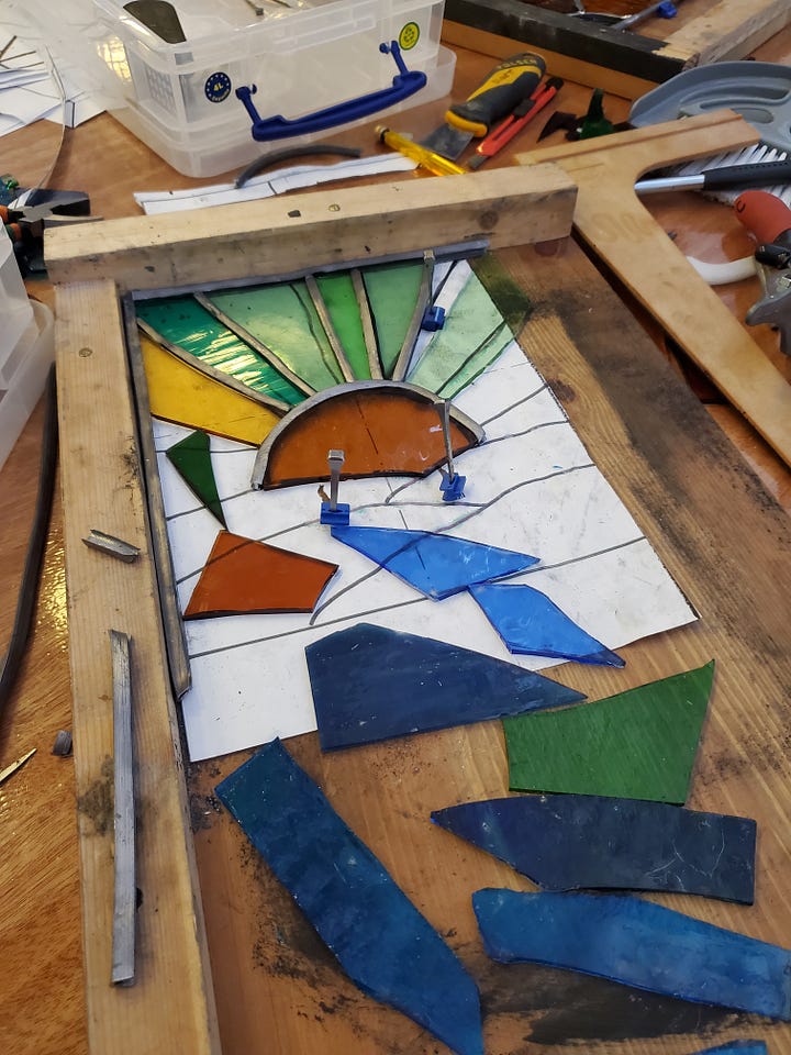 First Image: A person is working on a stained glass project. Their hands are carefully arranging pieces of colored glass, including yellow, gold, and black sections. Tools like pliers and a cutting tool are visible on the table, alongside various sheets of glass and a wooden frame. There is a paper design outline partially shown underneath the glass pieces. Second Image: A partially completed stained glass artwork is in progress, showing a colorful sunburst design. Green and yellow glass rays emanate from an orange half-sun, with blue and green glass pieces arranged at the bottom. The design is laid out in a wooden frame, with scattered glass pieces and tools like clamps, pliers, and a cutting tool visible around the workspace.