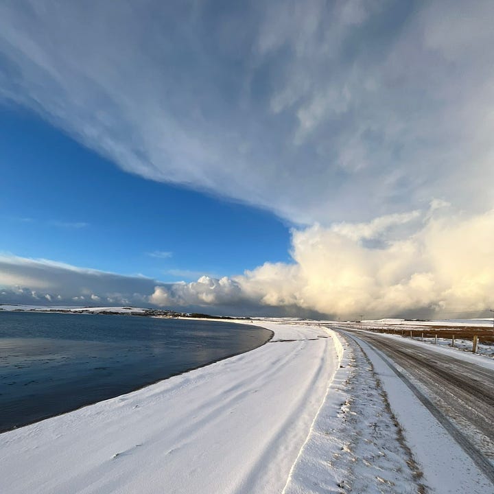 Top left: view across a field of heather with a dilapidated farm building in the foreground. Background is the blue sea & bluer sky. Top right, yellowy image taken on the beach on a drizzly day, a full rainbow sits on top of the sand & you can just make out a second rainbow above it. Bottom right - blurry image of the northern lights, green flashes on a dark background. Bottom left - image of the beach, sea on the left & shore on the right. the shore is covered in snow. The sky is blue with some cloud. 