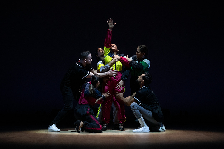 Two side-by-side images: On the left, against a red backdrop, three dancers in a V formation raise their arms to the sky, gazes lifted. They wear jumpsuits and flamenco shoes. On the right, against a black backdrop, one dancer reaches a palm upward as others gather around her, their hands resting on her torso and legs.