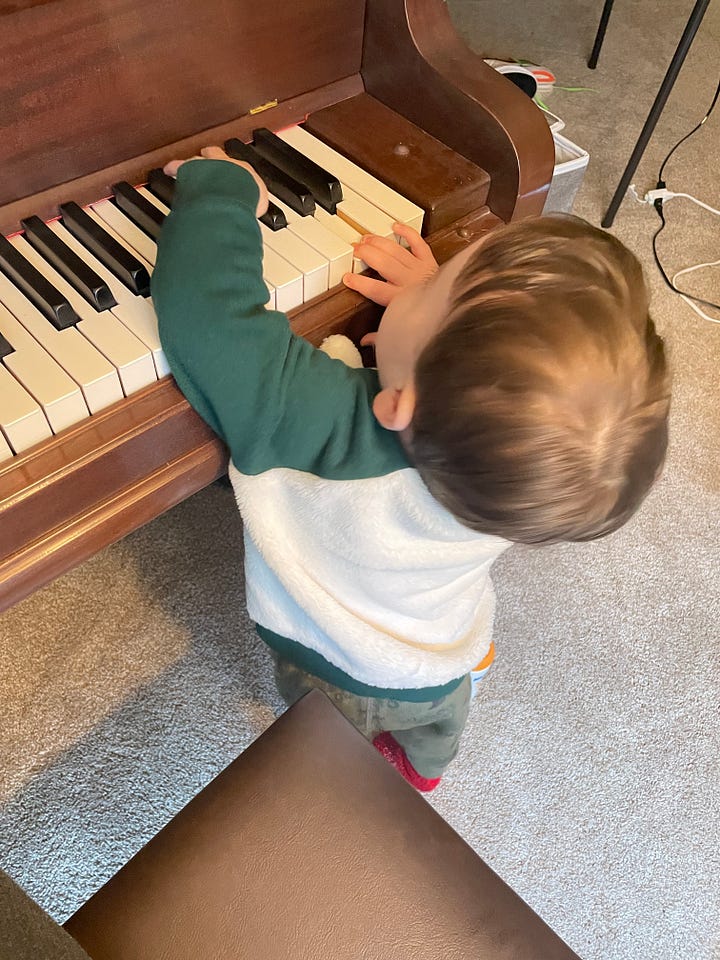 A baby grand piano stands in the corner of a room flooded with natural light. A toddler stands on the ground reaching up to touch the keys.