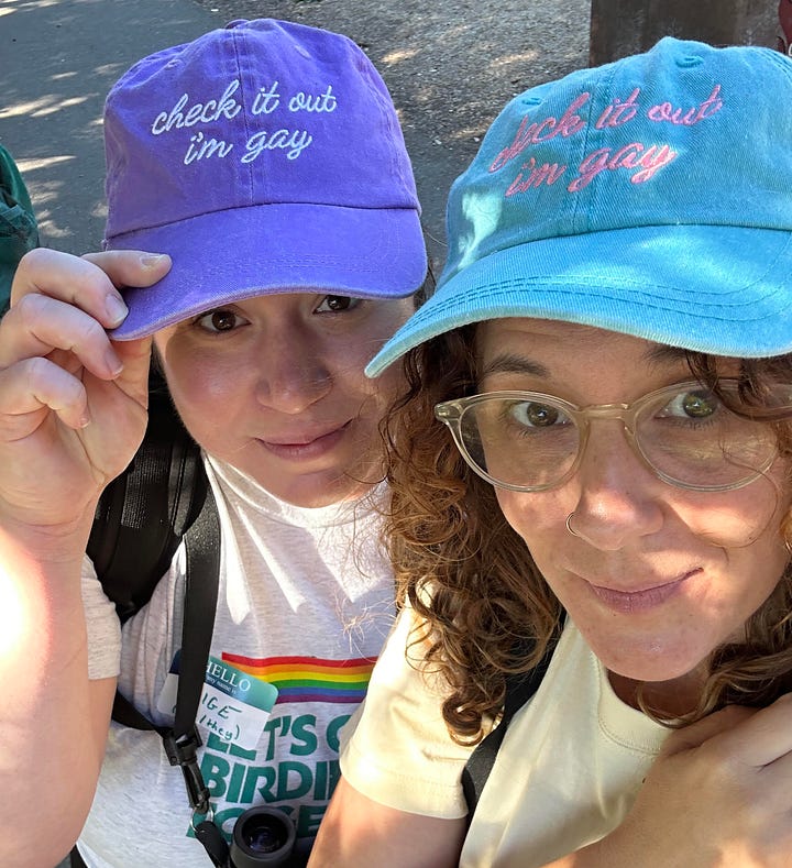 The left image shows QBBA co-leads Paige Pritchard and Alyssa Winn wearing purple and blue hats, cheekily embroidered with the phrase "Check it out, I'm gay." On the right, a selfie taken by Paige shows a group of seven birders on a hard-pack gravel trail through what appears to be a cedar or pine-filled wooded area.