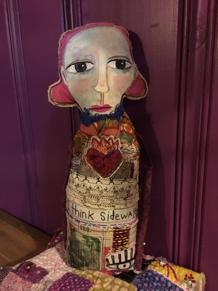 'Clockwise from top left, a plush doll with a brown face, a purple body and multicolored (primarily purple, pink and orange) feathers; the "not perfect" doll features a rabbit-eared figure with "not perfect" written over their mouth, they're sporting a yellow jumper with a big crimson heart on it; bottom, right, is a visual painting, "life," featuring two figures in Deveney's evocative "drip-like" style; and last, a doll with a bowling-pin-like body, with big sympathetic dark eyes, two big low ears that begin where earlobes would and a multicolored jumper on the body with knitted elements and a big red heart.