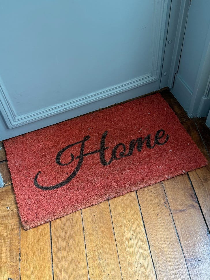 A red welcome mat has the word "Home" printed in elegant cursive. It sits on an old wood floor at the base of a light blue door.