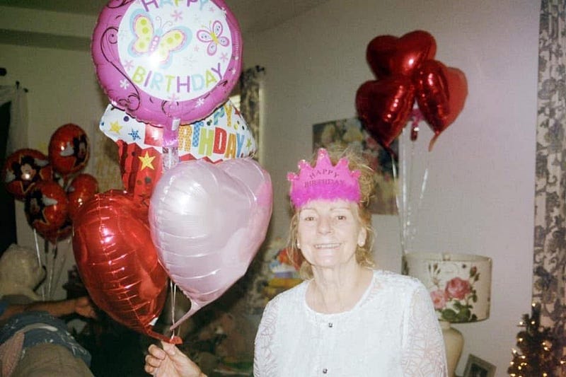 Kailee's Nana smiling in a pink paper crown, holding birthday balloons